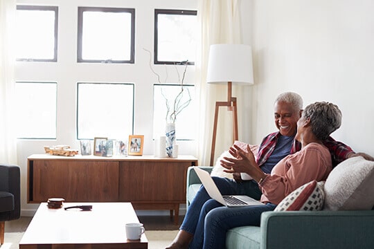 Senior woman holding laptop and talking to her husband while sitting together on sofa