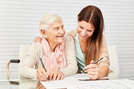 A female resident care provider sitting at a table with a senior resident helping with her with paper work