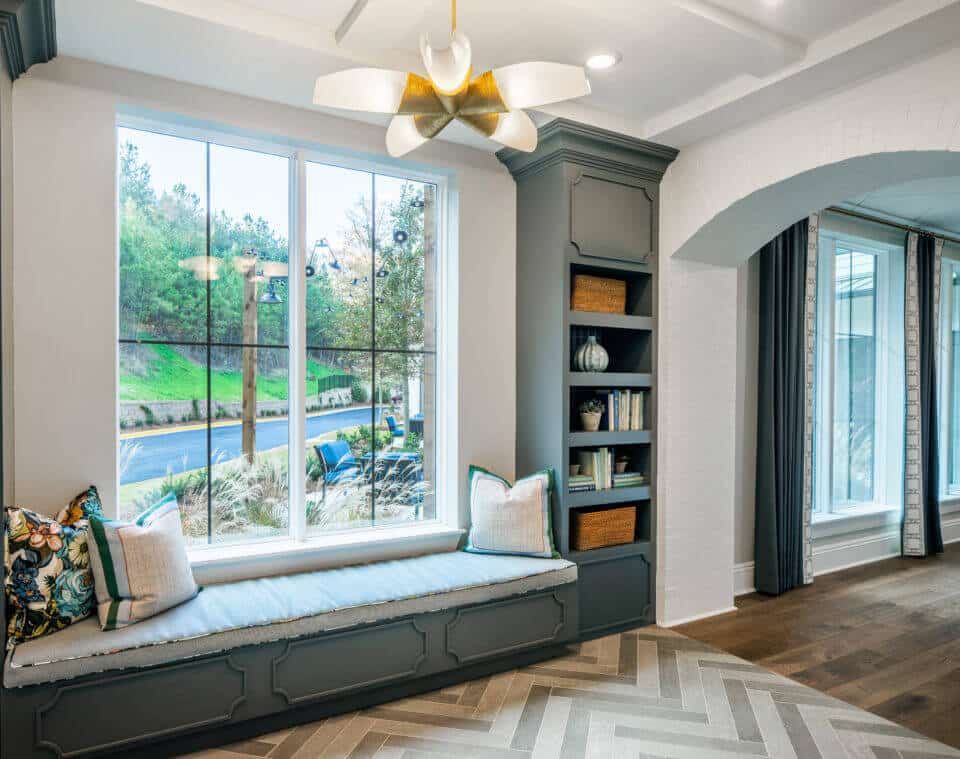 Longleaf common space window with bench setting and pillows, gray paneling and bookshelf, view of patio and green trees