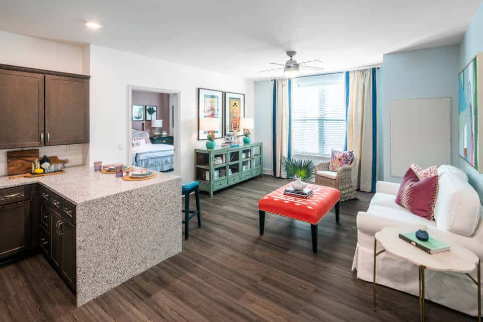 Living room white couch, red ottoman, green buffet table and wicker chair with kitchen bar to left and bedroom peeking through doorway in Longleaf assisted living model apartment