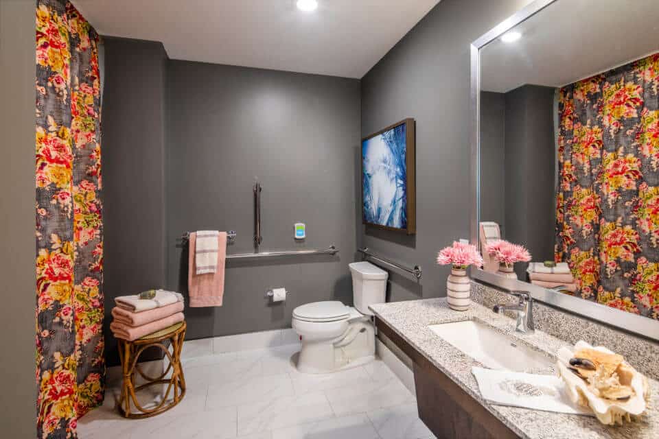 Bathroom vanity, toilet and shower with floral curtains, grey walls white floor in Longleaf assisted living model apartment