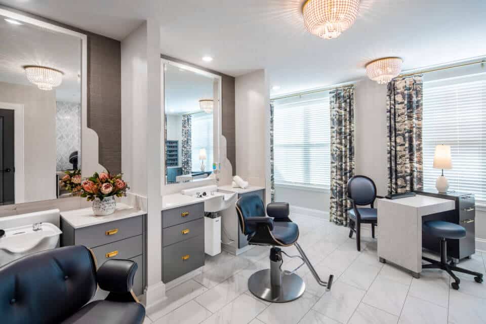 Two stylist stations with client chairs, white sinks and countertops, gray cabinets and mirrors with manicure table and chairs to right by windows in Longleaf salon