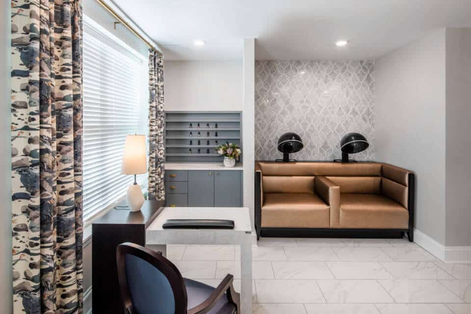 White manicure table with blue chair next to window with blue and white curtains at left and two bronze salon chairs with dryers at back