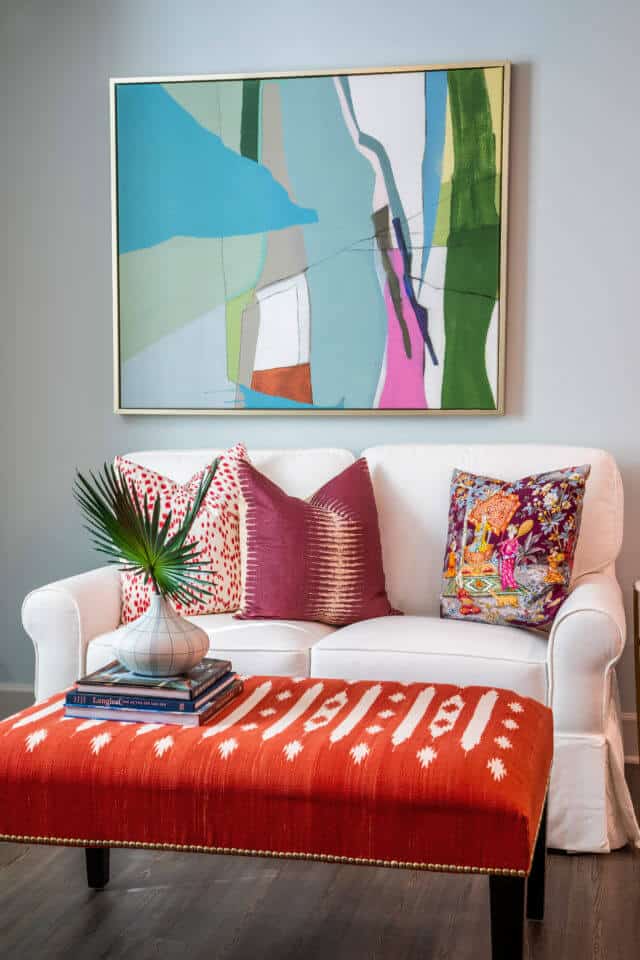 White living room sofa with colorful pillows, red ottoman, and painting on wall in Longleaf assisted living model apartment