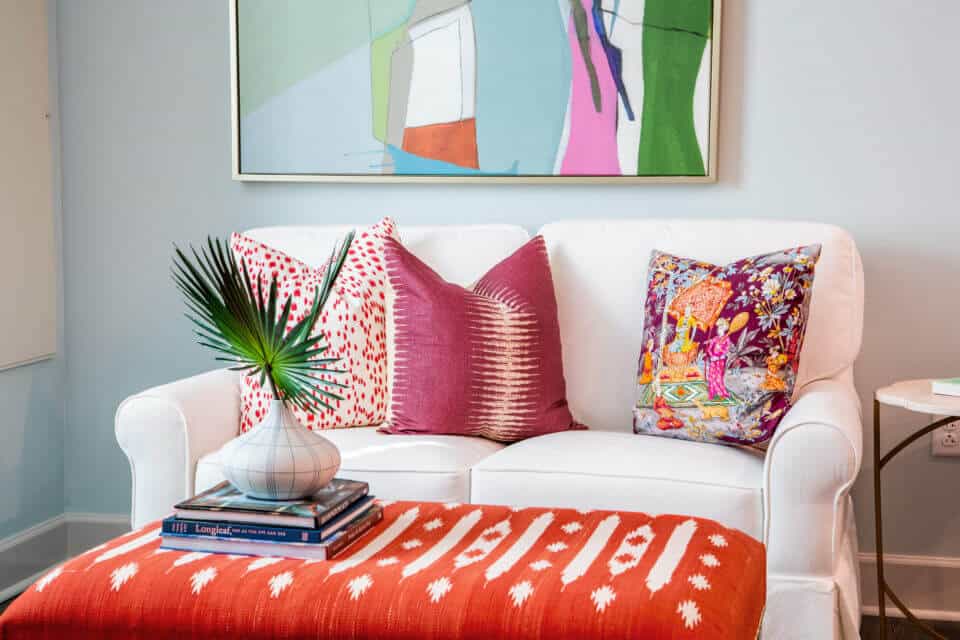 White living room sofa with colorful pillows, red ottoman, side table and painting on wall in Longleaf assisted living model apartment