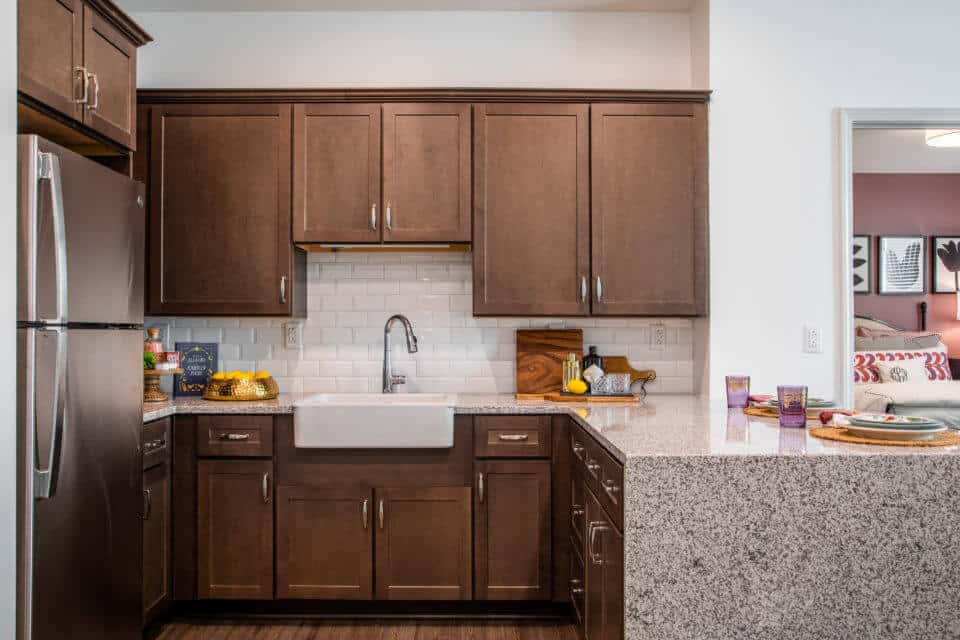 Kitchen with waterfall granite countertops, white farmhouse apron sink and brown cabinets with stainless steel refrigerator and bedroom peeking through doorway at right