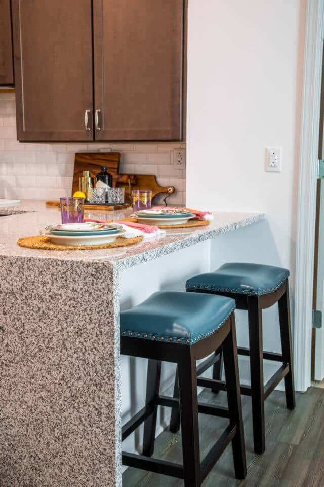 Two teal and brown stools at breakfast bar with waterfall granite countertops in kitchen with white wall, white backsplash and brown cabinets in Longleaf assisted living model apartment