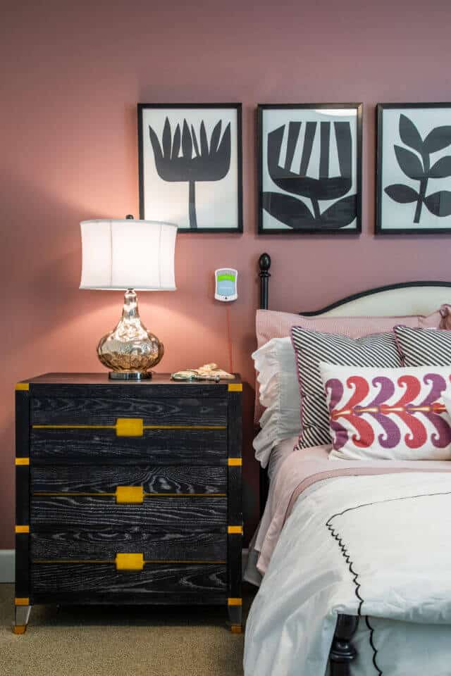 Left side view of white bed and colorful pillows with black and gold nightstand and glass lamp with white shade and black and white wall art on mauve wall in Longleaf assisted living model apartment
