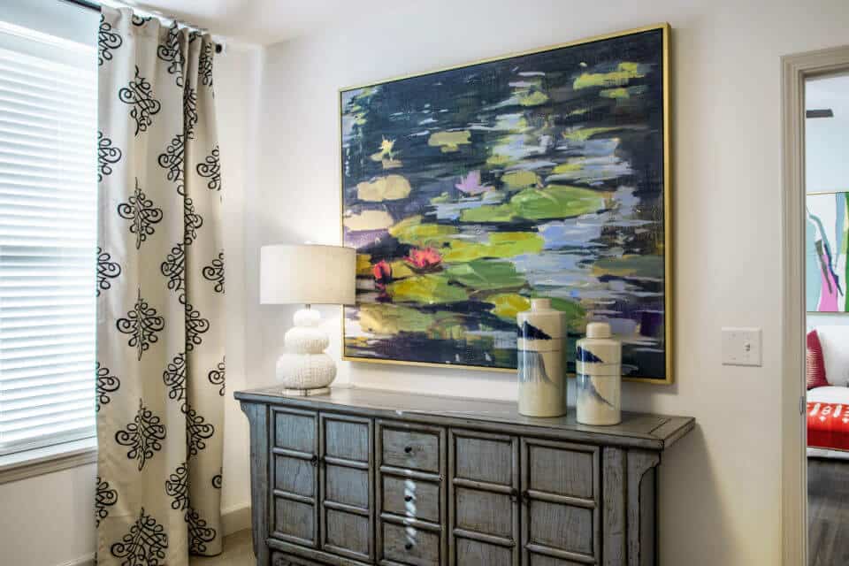 Grey distressed dresser with pottery, white lamp and blue/green painting on wall and window with navy and white curtains at left in Longleaf assisted living model apartment