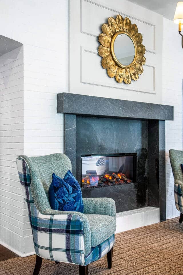 Black see through fireplace outlined by white brick wall and circular gold mirror hanging above with two green plaid chairs on each side in Longleaf common space