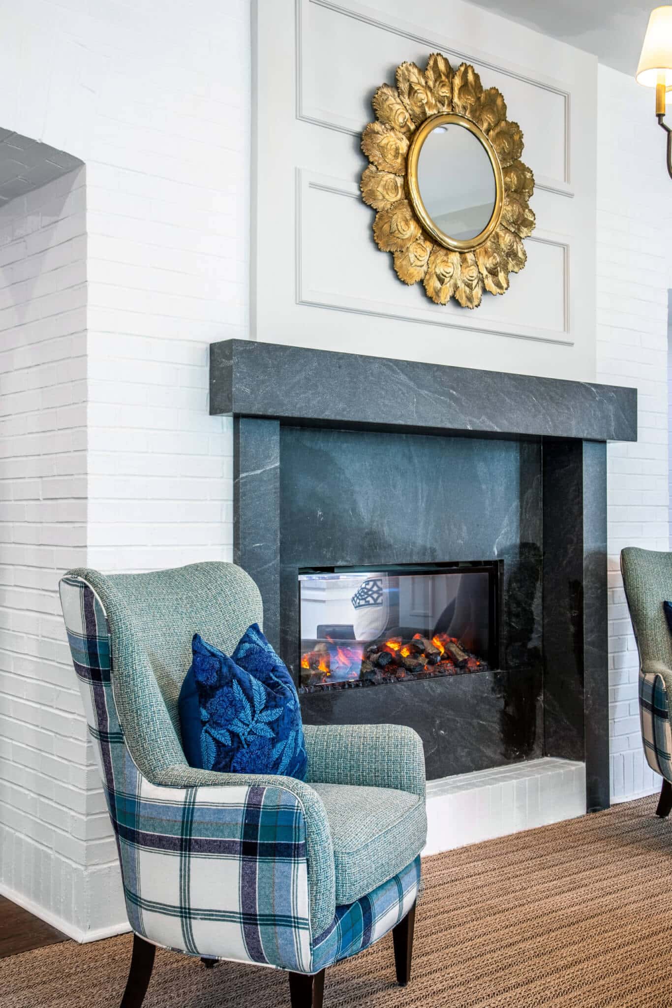 Black see through fireplace outlined by white brick wall and circular gold mirror hanging above with two green plaid chairs on each side in Longleaf common space
