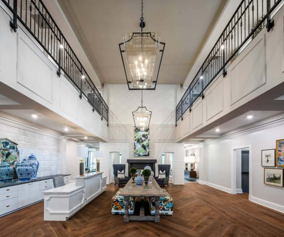 Longleaf foyer with white front desk at left, long wood table in center holding blue/white vases, greenery in pots and floral benches underneath, and open to second floor above with black metal railing, glass light fixtures, see-through fireplace at back of room with chairs