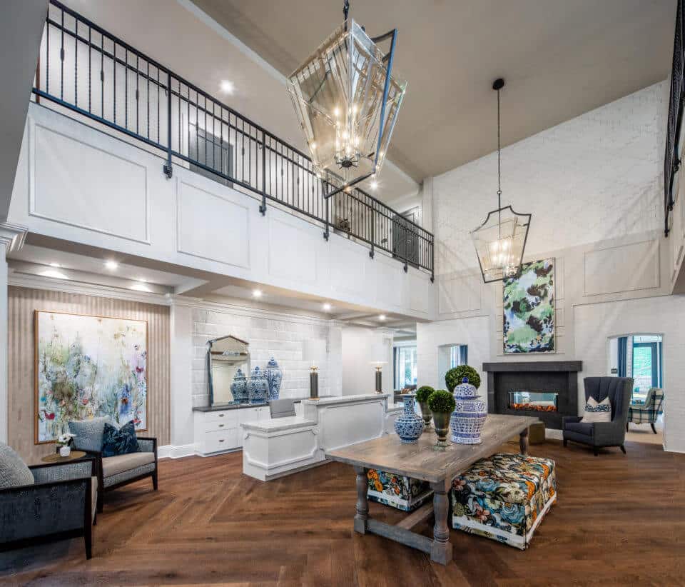 Longleaf foyer with chairs, table, painting, and white front desk at left, long wood table in center holding blue/white vases, greenery in pots and floral benches underneath, and open to second floor above with black metal railing, glass light fixtures, see-through fireplace at back of room with chairs