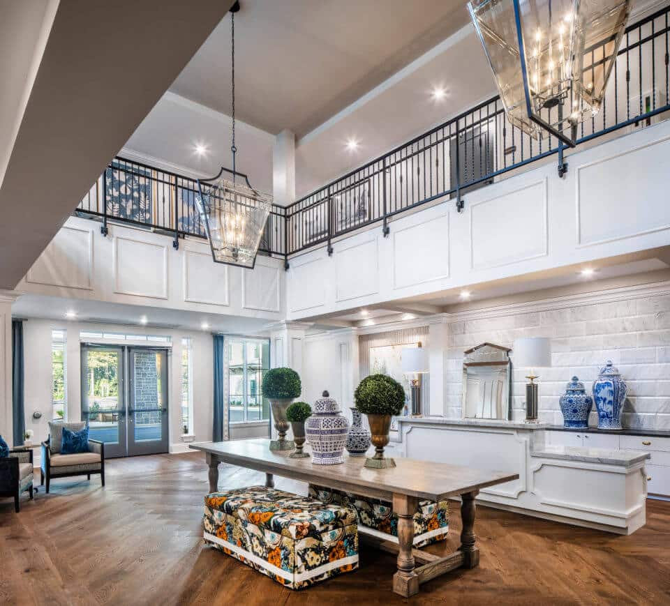 Longleaf foyer looking at entrance to left, white desk at right and long wood table in center holding blue/white vases, greenery in pots and floral benches underneath, and open to second floor above with black metal railing, glass light fixtures