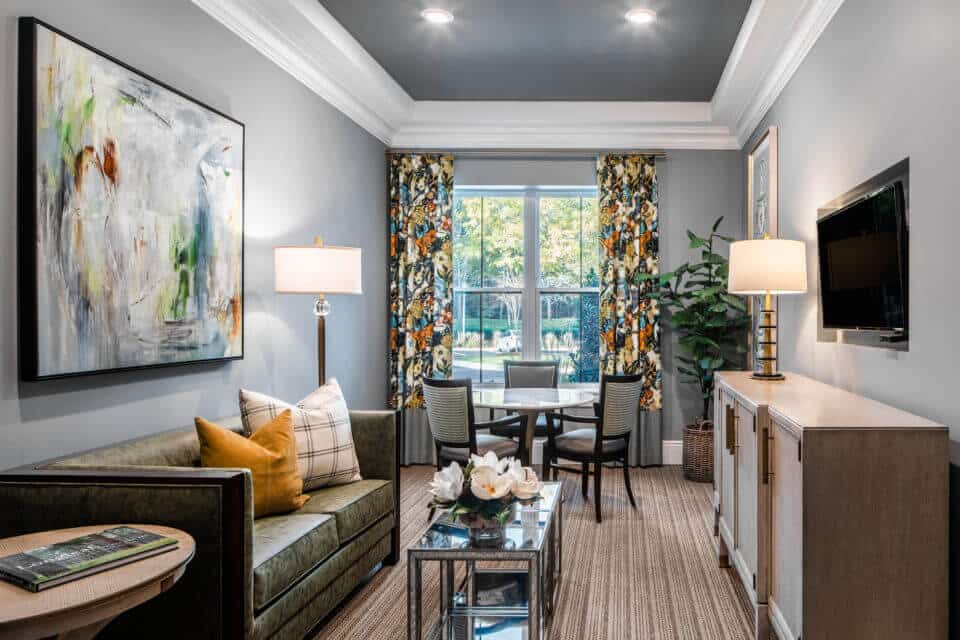 Longleaf discovery room with green sofa at left, glass coffee table in center and flat screen tv mounted in wall at right over wide dresser with round table and chairs in front of window at back of room with floral curtains