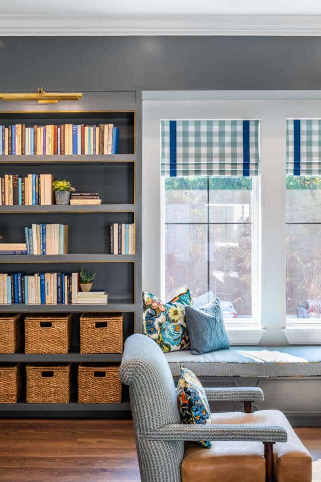 Longleaf library with window above bench seating, blue pattern and brown leather chair, gray wood bookcases with books and baskets on shelves