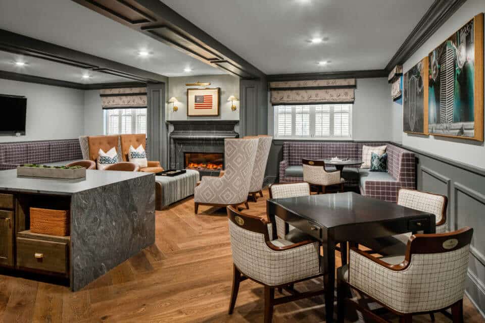 Longleaf sports lounge with bar at left, square table and four white plaid chairs at right, corner bench seating at back right, fireplace with four chairs and ottoman in center back and two windows on each side.