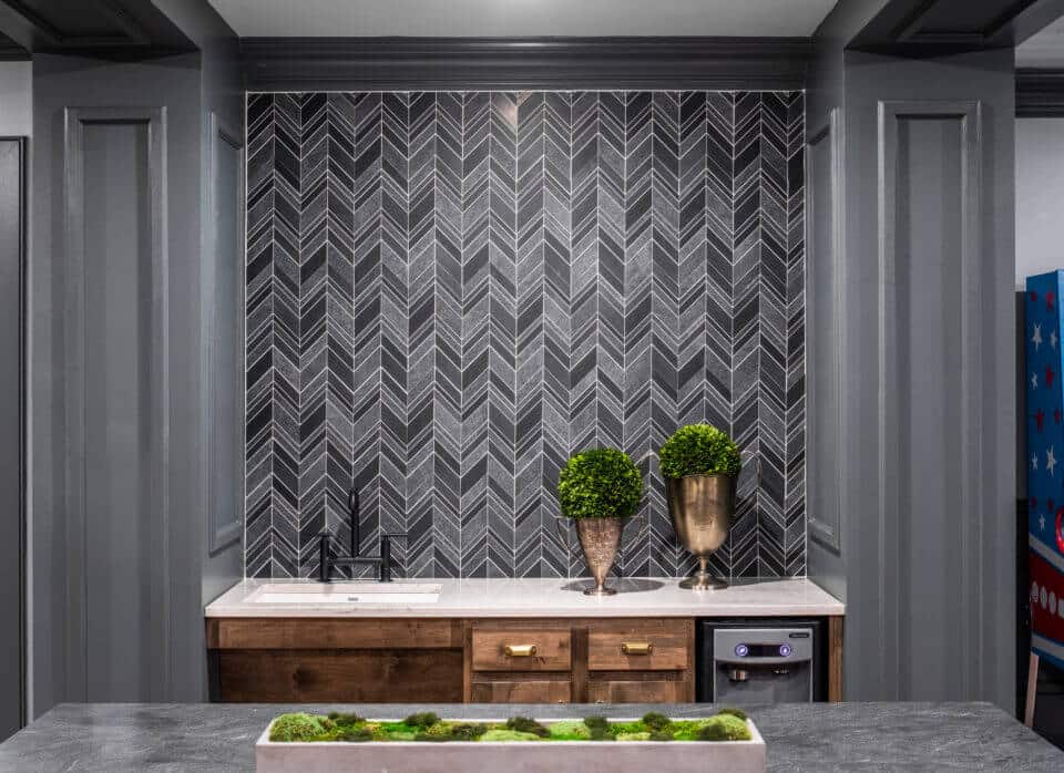 Longleaf lounge top of bar with gray chevron backsplash and gray walls, brown cabinets with white countertop and sink greenery in pots and vases