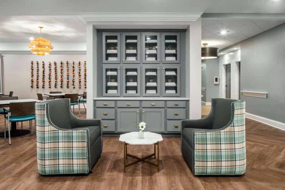 Longleaf common space with two green plaid chairs, white table and gray cabinet at back, tables and chairs at left