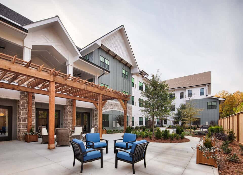 Longleaf patio view of pergola with wicker chairs and white cushions, blue chairs and wooden privacy fence at right