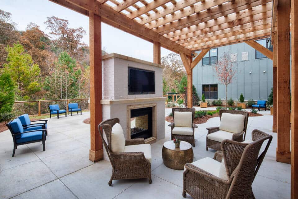 Longleaf patio under pergola with wicker chairs and white cushions, blue chairs and fence with wooded view