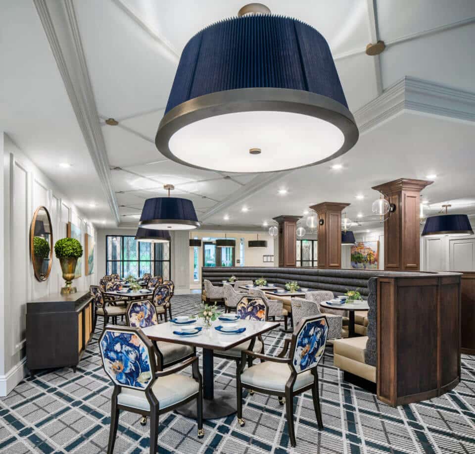 Longleaf main dining room with blue light fixtures, blue and beige curved booth, tables and floral backed chairs with wood columns and globe light fixtures at right, buffet table, mirror and potted plant at left