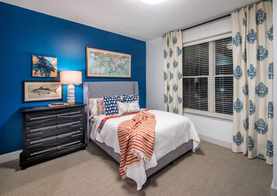 White bed with colorful throw and pillow, black nightstand with lamp and artwork above window and curtains at right in Longleaf memory care model apartment