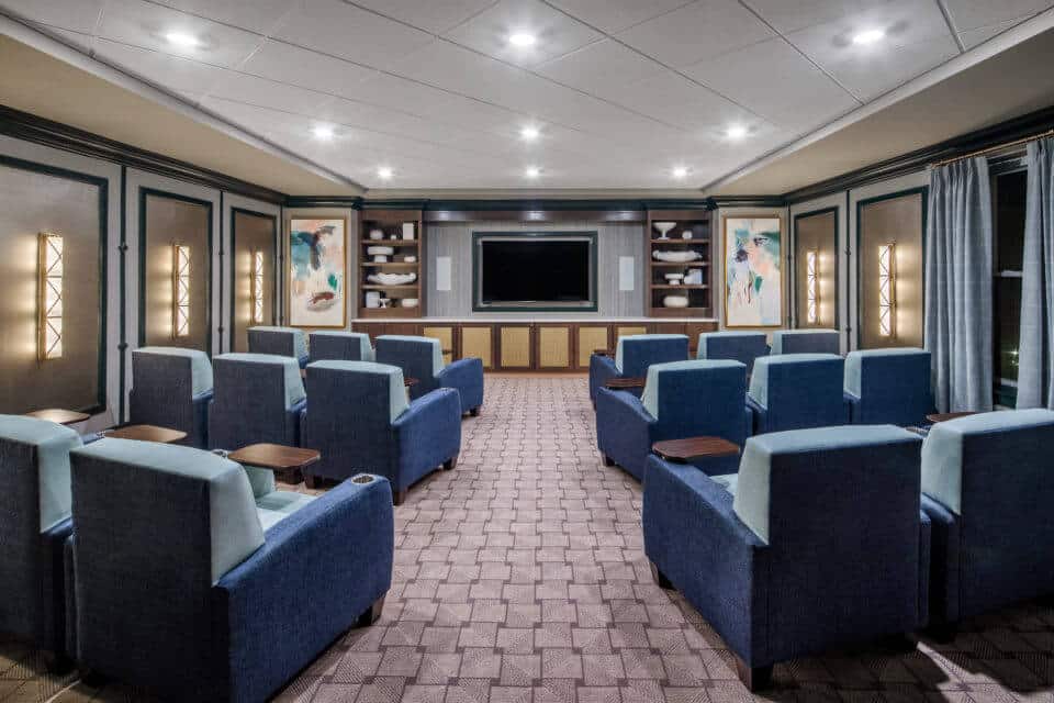 View from rear of Longleaf movie theater with blue seating, theater screen centered by wood shelves and paintings, side lighting