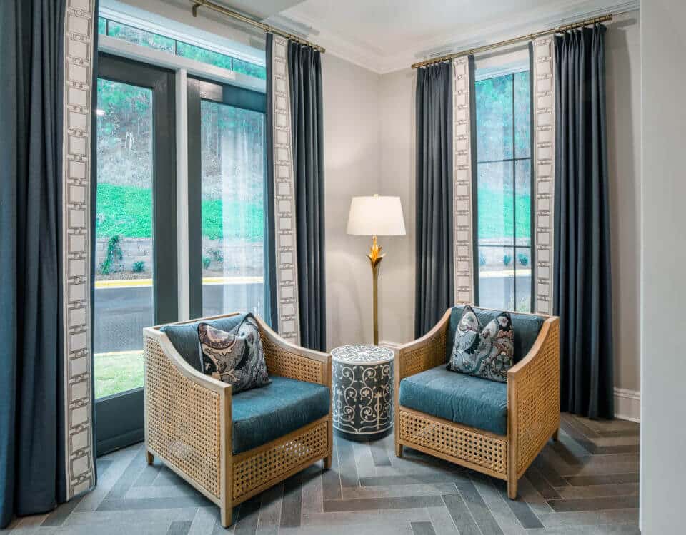 Two wicker chairs with blue cushions, table and lamp, in front of windows with blue gray curtains in Longleaf common space