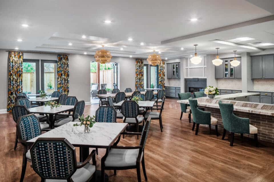 Longleaf memory care dining room with four person tables at left, long table at back, bar with green chairs and grey cabinets at right, three white light fixtures hanging above bar and two basket weave light fixtures above long table, floor curtains on windows at back