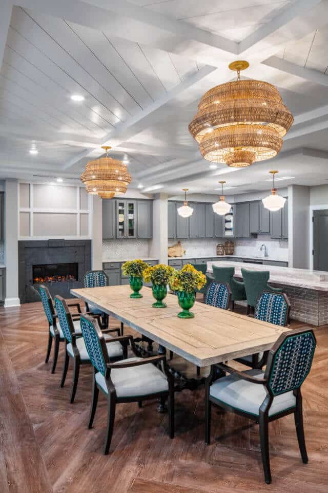 Longleaf memory care dining room long table with chairs, basket weave light fixtures, fireplace at back, bar with three green chairs and grey cabinets at right, three white light fixtures hanging above bar