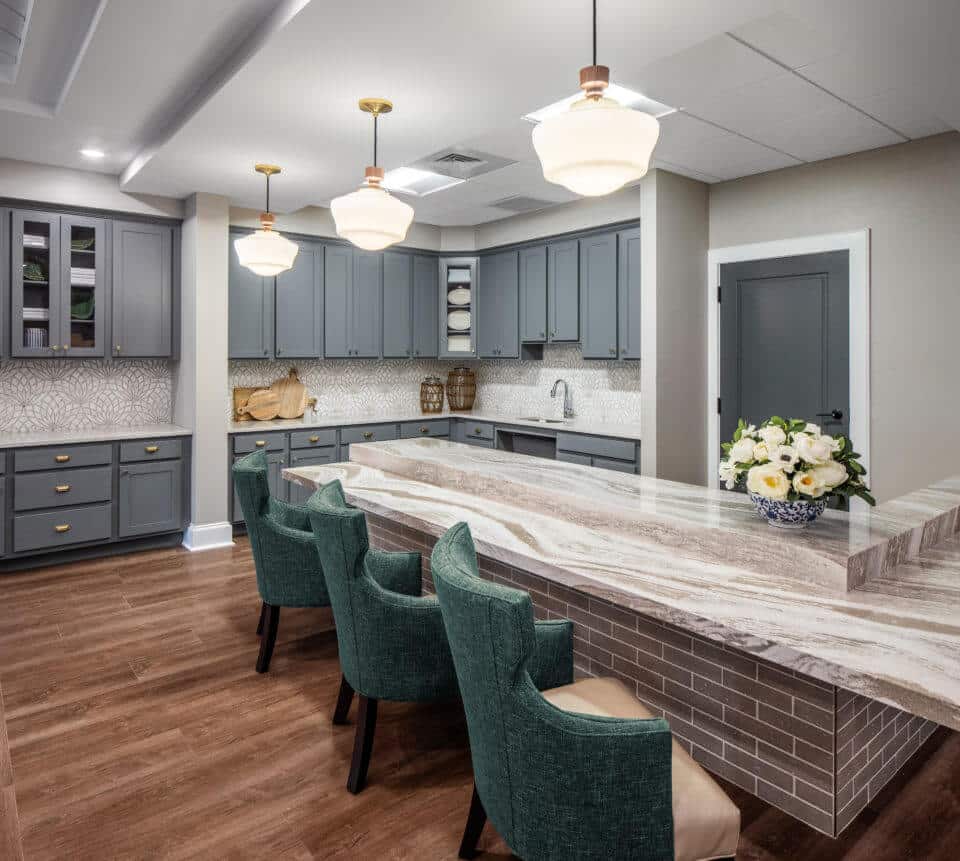 Longleaf memory care dining room bar with three green chairs and grey cabinets at back, three white light fixtures hanging