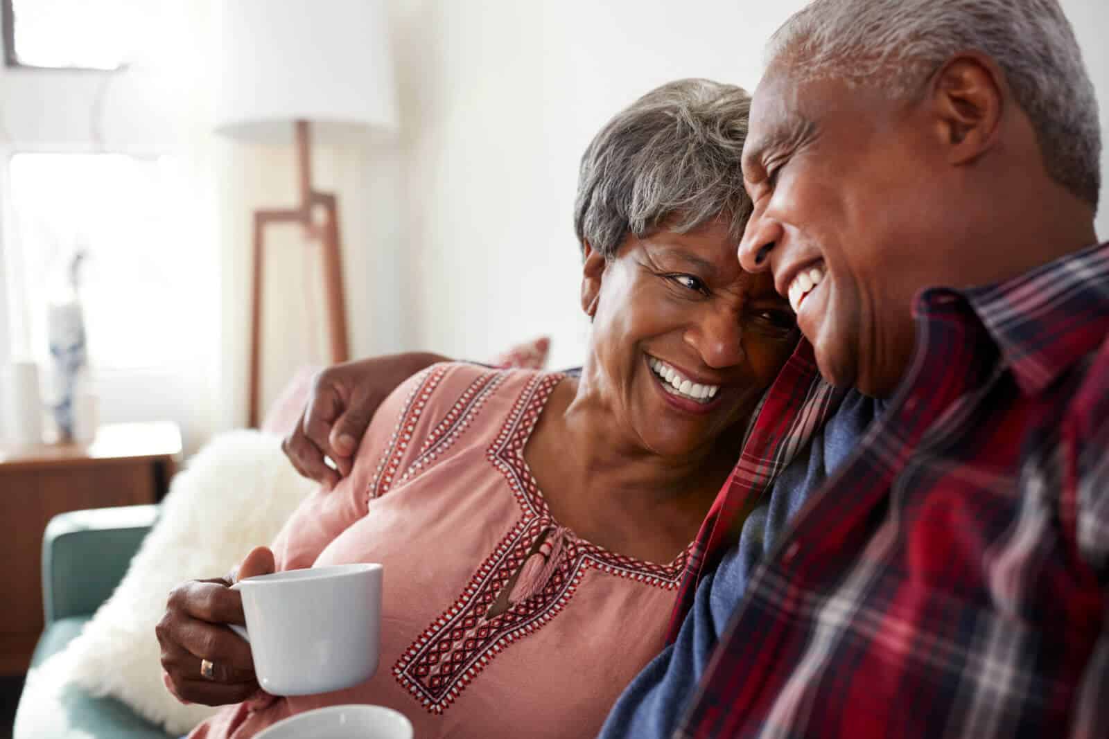 Senior couple sitting together in embrace on sofa smiling and holding white coffee mugs